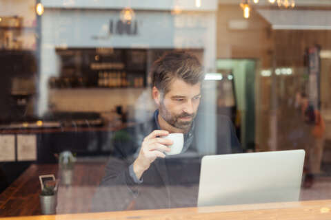 Work-Life-Balance: Ein Mann sitzt hinter einem Café-Fenster und arbeitet am Laptop. Er trinkt dabei Kaffee. Darstellung Work-Life-Balance