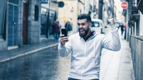 WM-Apps für den Fußball-Sommer: Junger Mann mit Bart schaut auf der Straße auf sein Smartphone und jubelt.