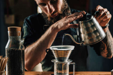 Filterkafffee: Ein Mann gießt Wasser aus einer Edelstahlkanne in ein Glas mit Filteraufsatz. Der Kaffee läuft durch.