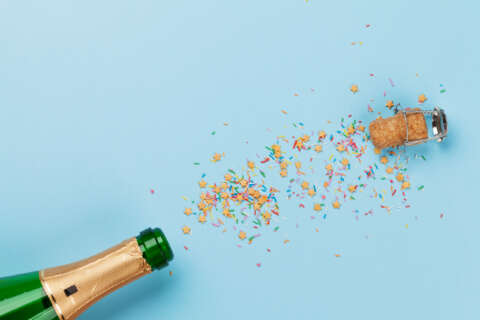 Alkohol: Sektflasche vor blauem Hintergrund. Korken und Konfetti fliegen aus der Flasche