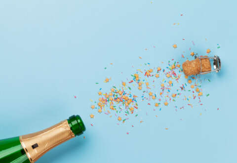 Alkohol: Sektflasche vor blauem Hintergrund. Korken und Konfetti fliegen aus der Flasche